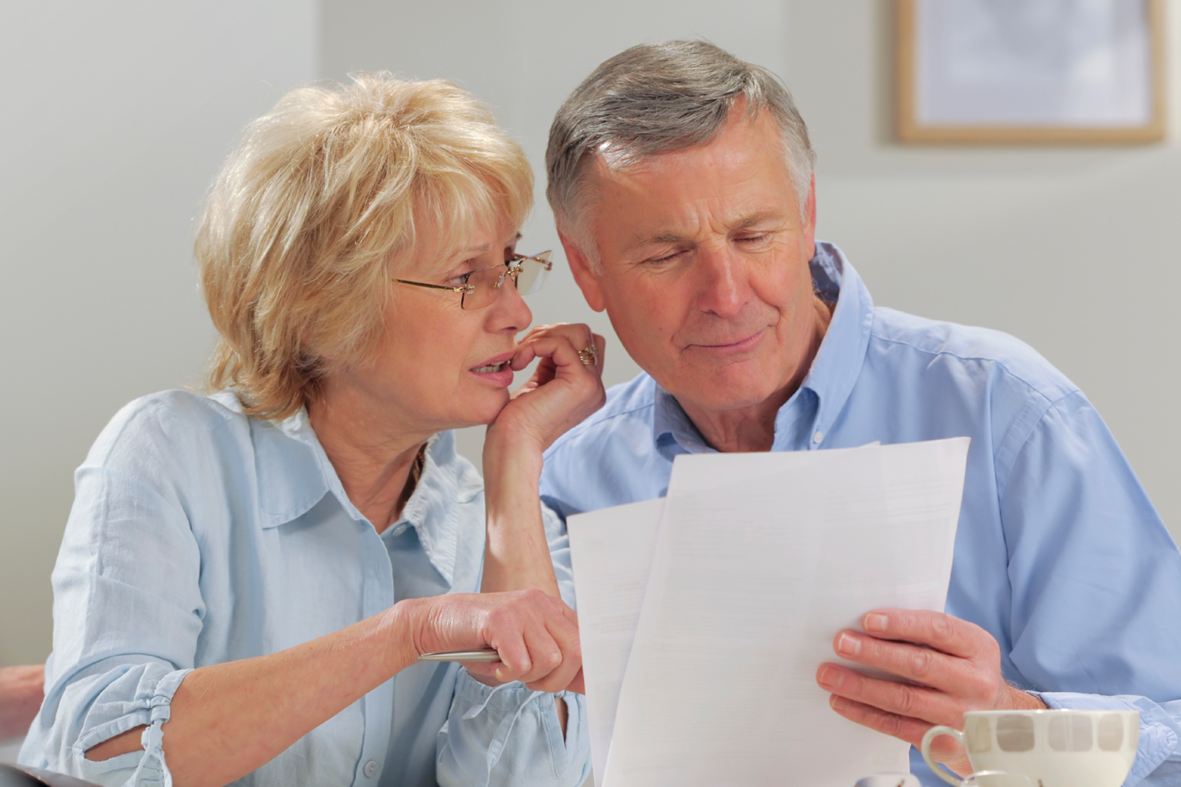 An older couple sits together reviewing documents at a table, both appearing focused. The woman leans in and points at the papers while the man holds them.