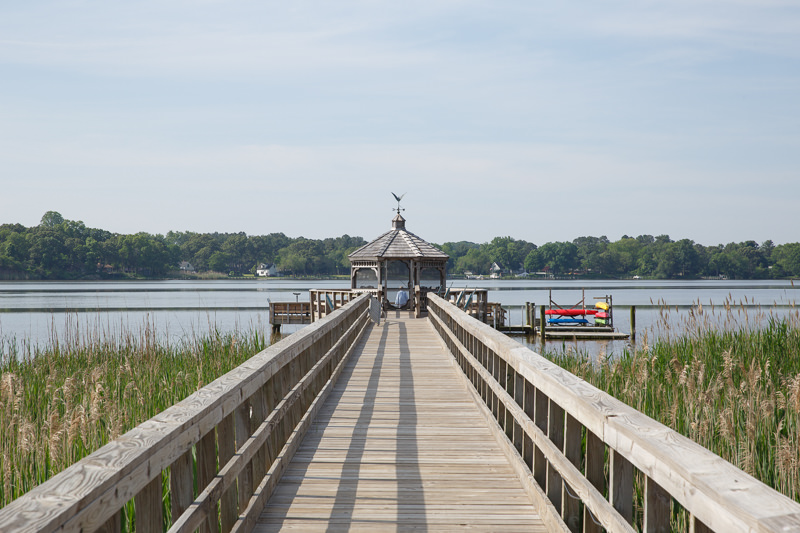 A wooden pier leads to a small gazebo over a calm lake, with reeds in the foreground and trees lining the far shore. A dock with kayaks is visible to the right.