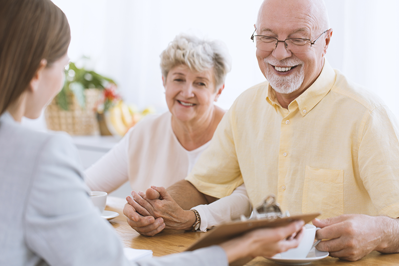 An elderly couple sits at a table, smiling and holding hands, while a woman across from them holds a clipboard and speaks to them.