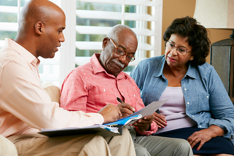 Three adults sit together in a living room reviewing documents; one man gestures toward the papers while the older man writes and the woman looks on attentively.