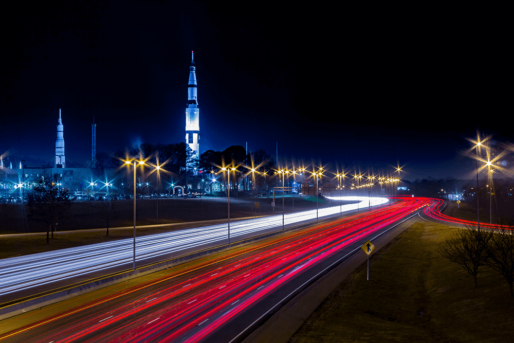 Night view of a highway with red and white light trails from cars; two tall rockets are lit up and visible in the background under a dark sky.