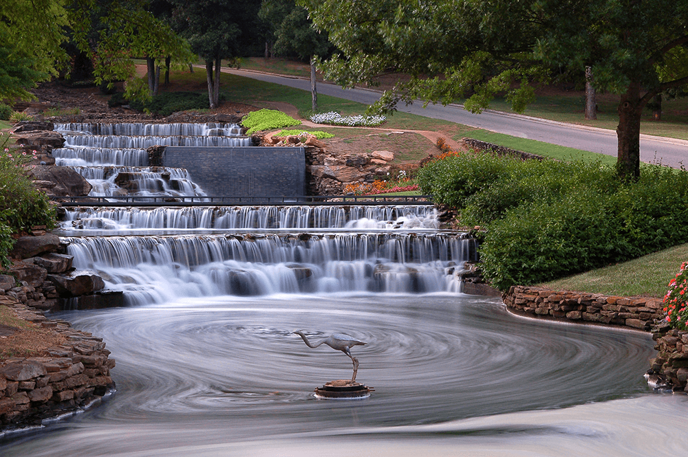 Terraced waterfalls flow into a swirling pool with a crane statue at the center, surrounded by greenery and a road in the background.