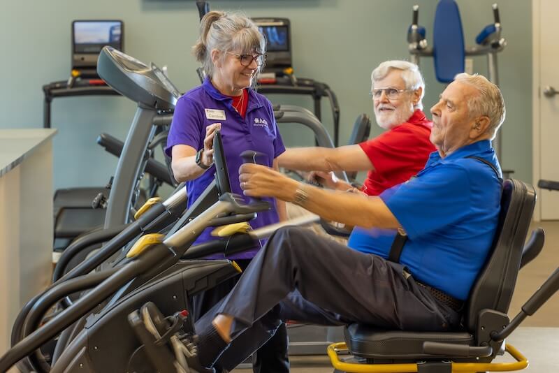 Two older men use exercise machines at a gym while a woman in a purple shirt stands nearby, smiling and assisting them.