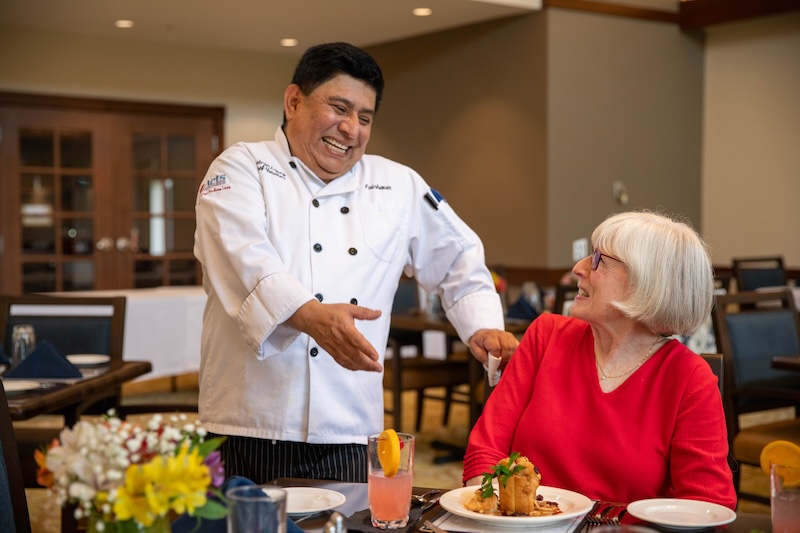 A chef in a white uniform gestures and smiles at a woman in red seated at a table with plated food and a pink drink in a restaurant setting.