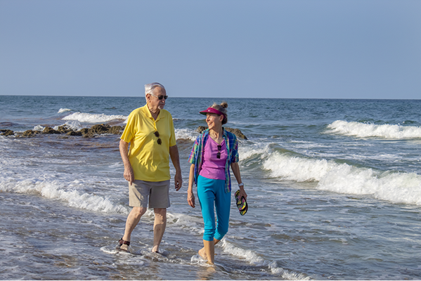 An older man and a younger woman walk barefoot along the shoreline, talking and smiling, with waves and a clear blue sky in the background.