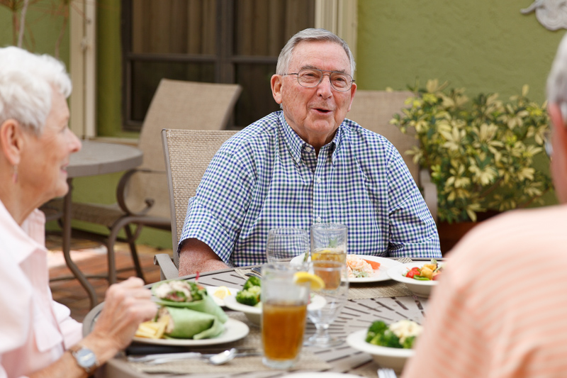 Three older adults sit at an outdoor table, eating a meal and drinking iced tea, with plates of food and glasses visible.