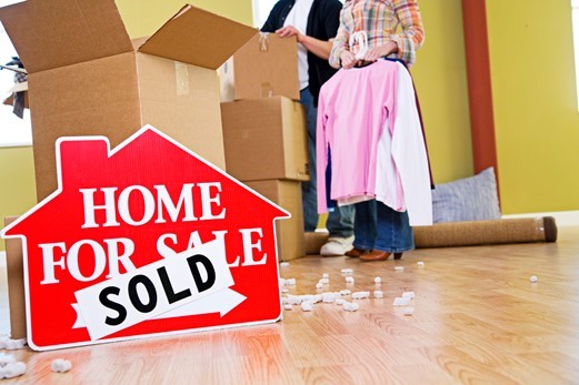 A red “Home For Sale” sign with a “Sold” sticker is in front of people packing boxes and clothes in an empty room with wood flooring.