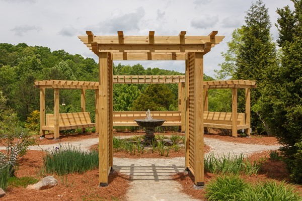 Wooden pergola structure with a curved bench surrounds a small tiered fountain, set in a landscaped garden with trees and mulched paths.