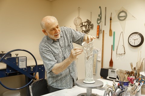 An older man sculpting a tall, intricate ceramic piece in a studio surrounded by pottery tools, brushes, and wall-mounted art supplies.