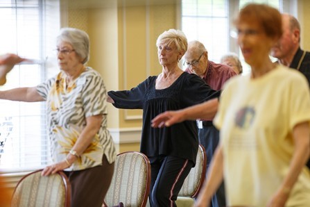 A group of older adults participate in a standing exercise class, with arms raised, in a well-lit room with chairs.