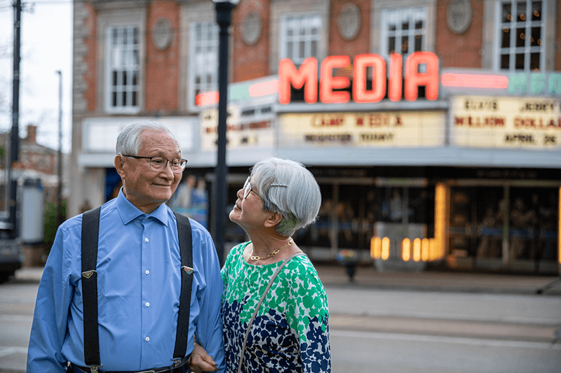 An older couple stands outside a theater with “MEDIA” on the marquee. The man wears a blue shirt and suspenders; the woman wears glasses and a patterned dress.