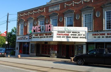 Brick theater building with marquee signs advertising “Footloose” summer musical, registration info, and subscriptions; cars parked in front along the street.