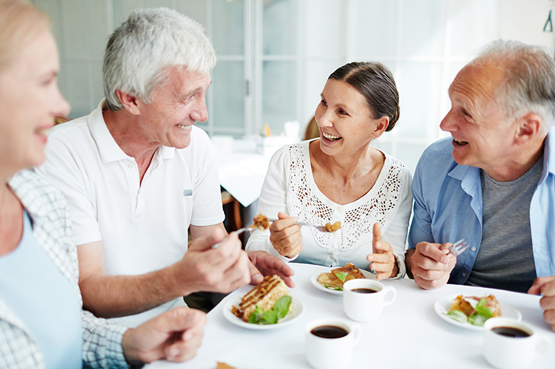 Four older adults sit at a table, eating food and drinking coffee while smiling and talking to each other in a bright indoor setting.