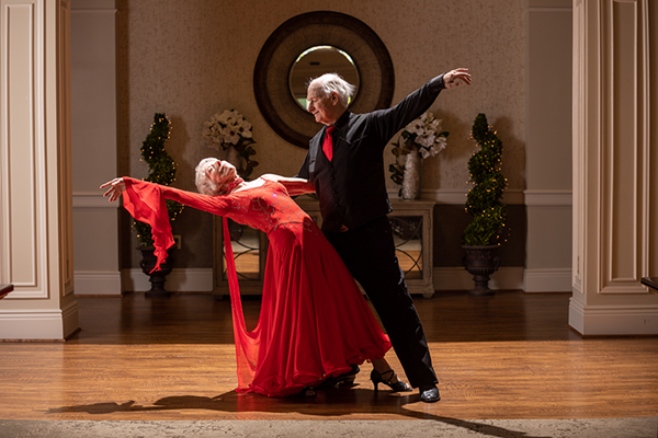 An older couple in formal attire performs a ballroom dance pose in a decorated indoor setting with wooden floors and potted plants.