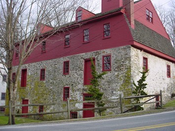 Three-story historic stone building with red upper level, red window frames, and a shingled roof, located beside a road with a wooden fence and leafless trees in the background.