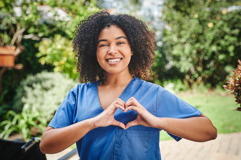 Smiling woman in blue scrubs stands outdoors, making a heart shape with her hands.