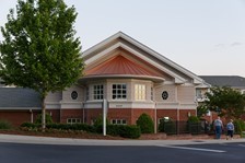 A brick and beige building with a triangular roof, round windows, and surrounding greenery. Two people walk on the sidewalk nearby.