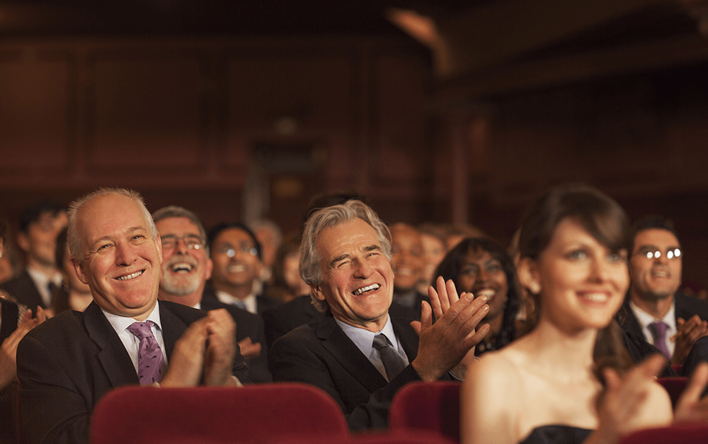 Audience members in formal attire sit in a theater, smiling and applauding. The background is dimly lit, creating a warm atmosphere.