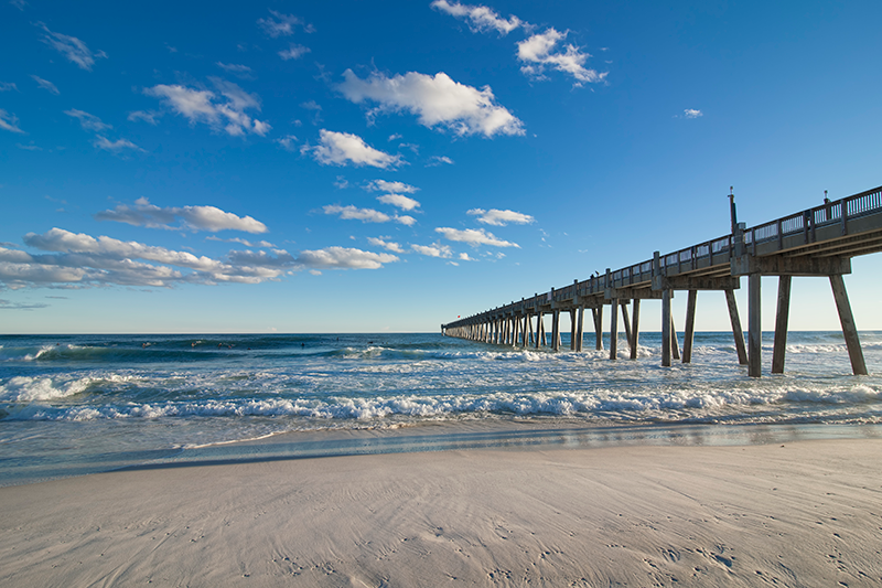 A long wooden pier extends over waves into the ocean under a blue sky with scattered clouds, viewed from a sandy beach.