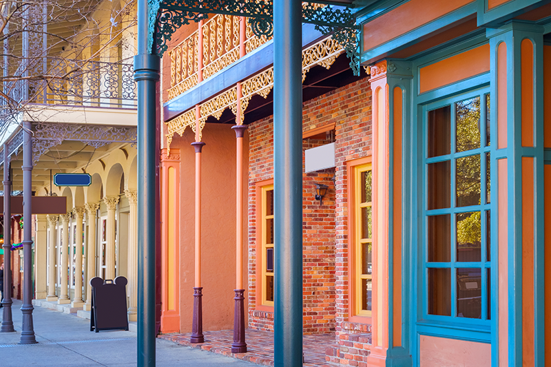 Colorful historic buildings with ornate ironwork, large windows, and brick facades line a quiet street under clear daylight.