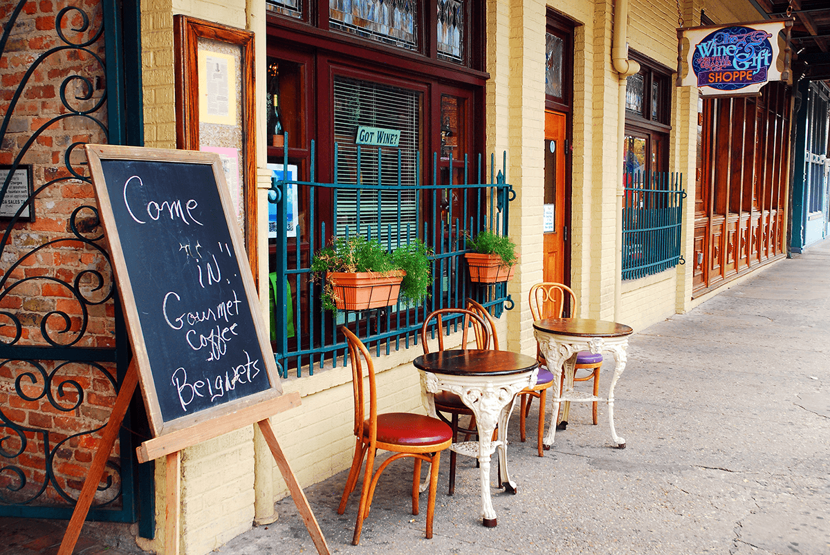 Two small tables with chairs are set outside a café with potted plants. A chalkboard sign advertises gourmet coffee and beignets. The window displays a “Got Wine?” sign.