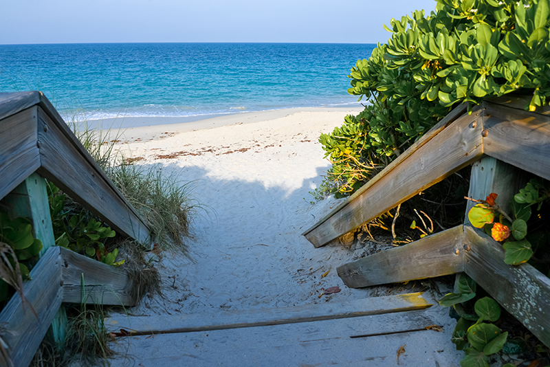 Wooden steps lead down to a sandy beach with green bushes on the side and a clear blue ocean under a bright sky.