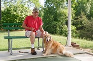 Man in red shirt and cap sits on a bench holding a leash, with a golden retriever lying beside him on a concrete patio, surrounded by grass and trees.