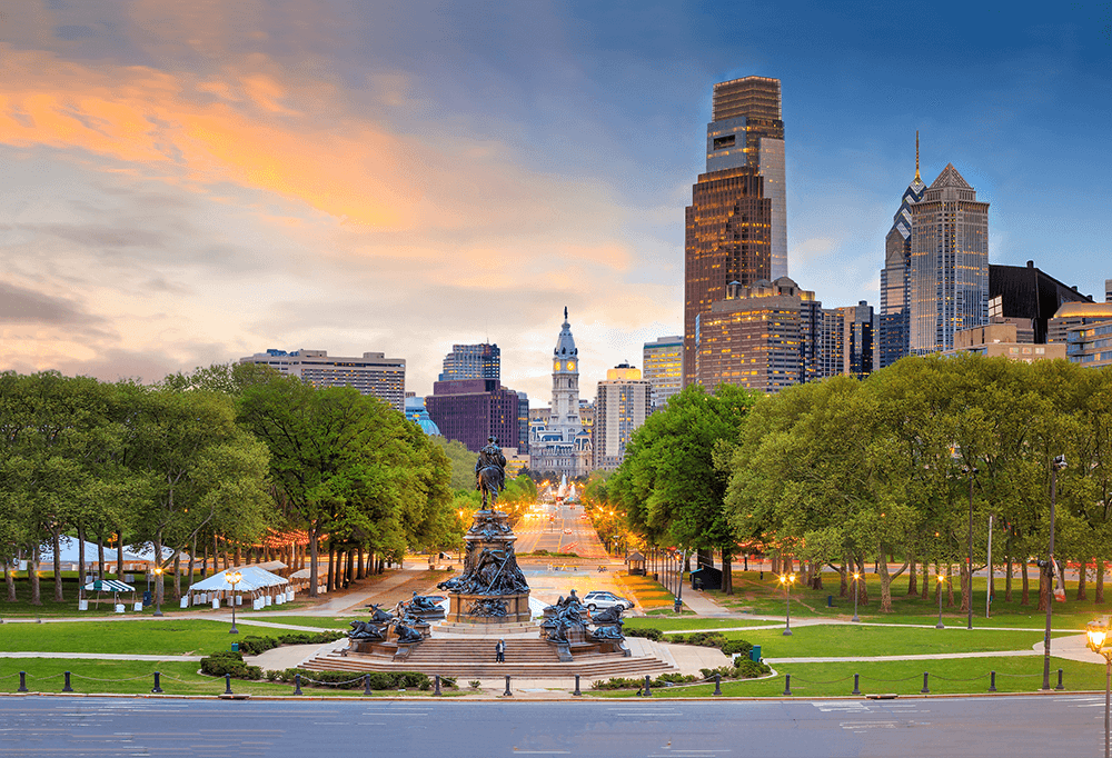 View of a city skyline with tall buildings, a central avenue, and a large monument in the foreground surrounded by green trees at sunset.