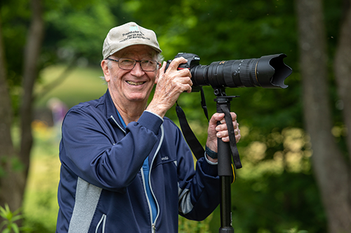 Older man wearing glasses and a cap stands outdoors, smiling while holding a camera with a large telephoto lens mounted on a tripod. Trees and greenery are visible in the background.