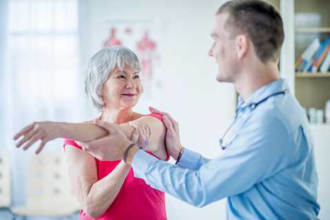 An older woman in a pink shirt has her arm gently stretched by a healthcare professional in a medical office.