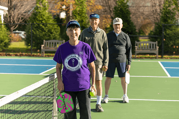 Three older adults stand on an outdoor pickleball court holding paddles, with the front person wearing a purple shirt and smiling at the camera.