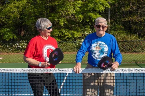 Two older adults stand on a pickleball court, smiling and holding paddles, with a green landscape in the background.