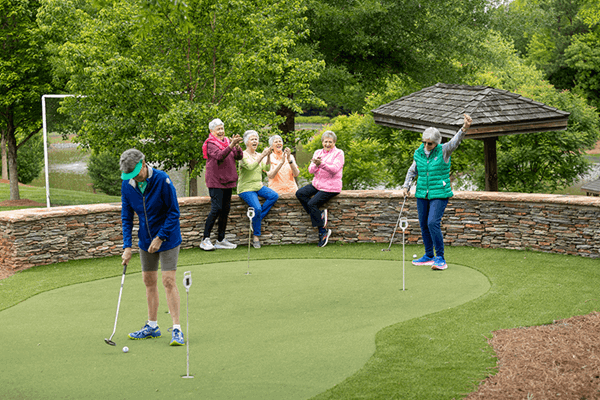 Six older adults enjoy a game of putting on a small green outdoors; two play while four others stand nearby, smiling and clapping. Trees and a gazebo are in the background.