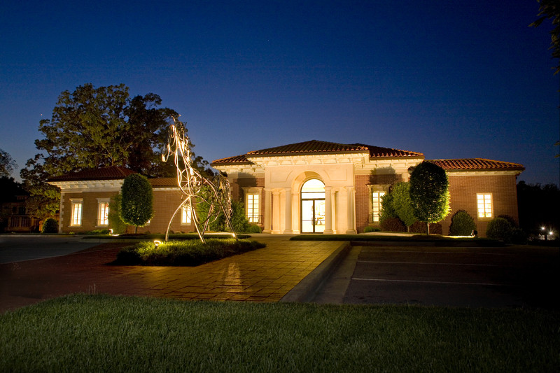 Single-story brick building with lit windows and arched entrance at night, surrounded by neatly trimmed shrubs and a metal sculpture in the front.