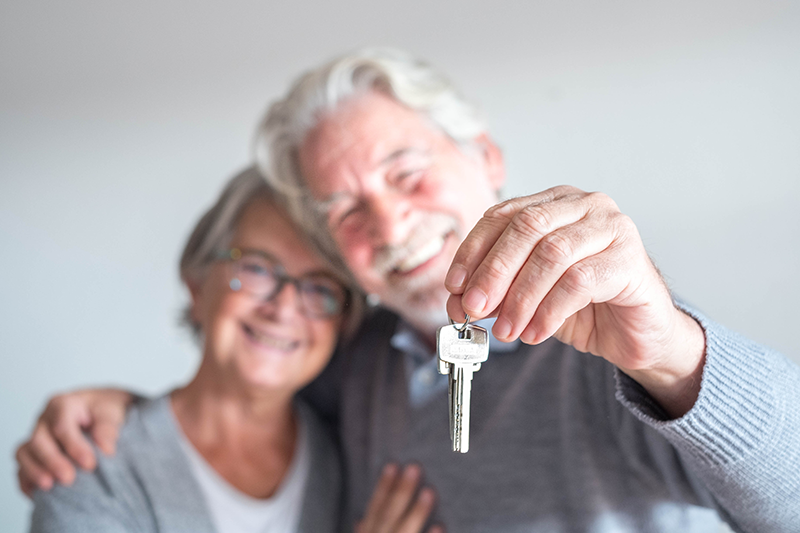 Smiling older couple standing together, with one person holding a set of keys towards the camera.