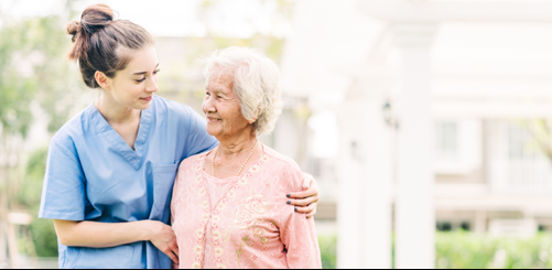 A young caregiver in blue scrubs stands outdoors with an older woman in a pink outfit, smiling at each other.