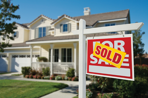 A “For Sale” sign with a “Sold” sticker stands in front of a two-story suburban house with a porch and yard.