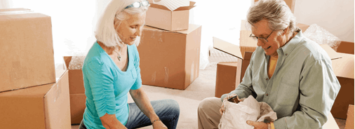 Two older adults unpack boxes in a bright room, surrounded by cardboard moving boxes and packing materials.