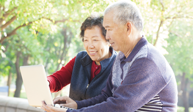 Two older adults sit outdoors, looking at a laptop together. The background shows trees and sunlight.