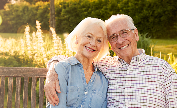 An elderly couple sits closely together on a wooden bench outdoors, smiling at the camera, with greenery and sunlight in the background.