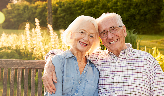 An elderly couple sits closely together on a wooden bench outdoors, smiling at the camera, with greenery and sunlight in the background.