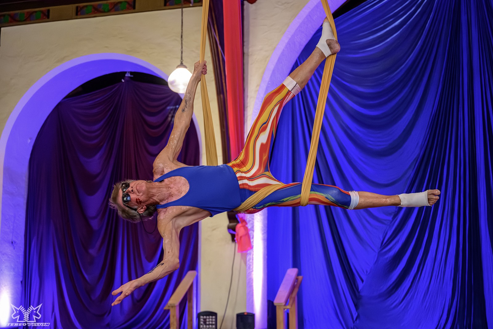 A male aerialist in a blue leotard and striped leggings performs a split pose on suspended fabric, with purple drapes and arched walls in the background.