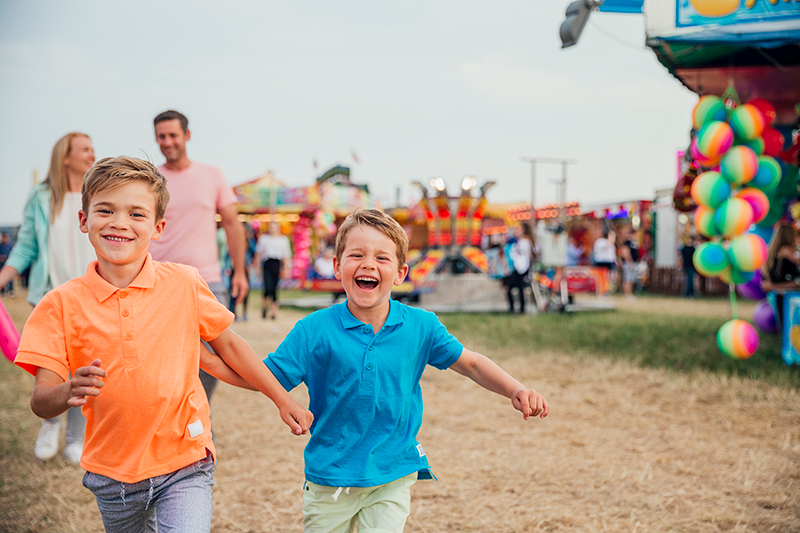 Two young boys holding hands and smiling as they run at an outdoor fair, with colorful rides, balloons, and other people in the background.