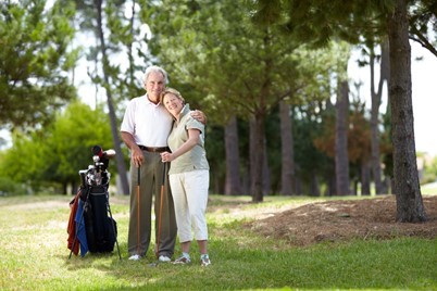 An older couple stands together on a golf course beside a golf bag, surrounded by trees and greenery, both smiling at the camera.