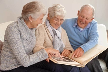 Three elderly people sit on a couch looking through a photo album together, smiling and pointing at pictures.