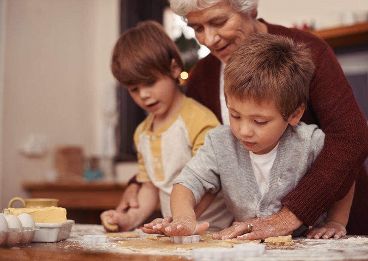 An older woman helps two young boys cut cookie shapes from dough on a floured kitchen counter.