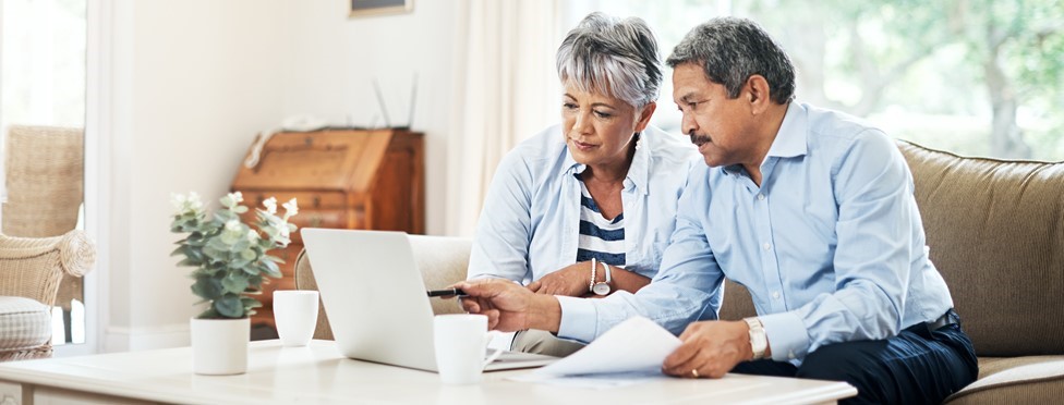 Two older adults sit on a couch, looking at a laptop screen and holding papers, appearing focused while working together in a bright living room.