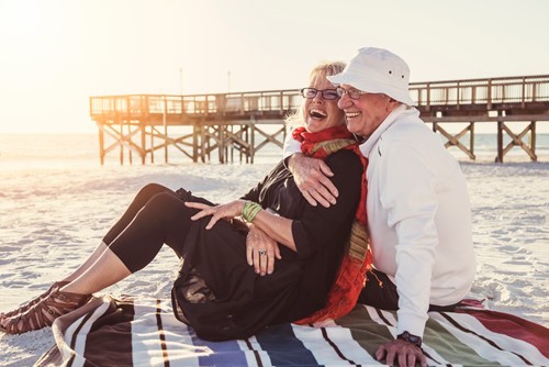 An older couple sits on a blanket at the beach, smiling and embracing with a pier in the background on a sunny day.