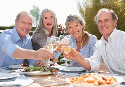 Four smiling adults sit at an outdoor table, raising glasses of white wine for a toast, with plates of food in front of them.