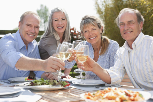 Four older adults sit outdoors at a table, smiling and raising glasses of white wine for a toast. Plates of food are on the table, and it is sunny outside.